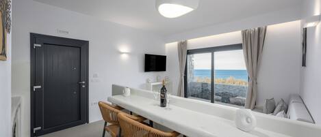 A modern kitchen with a long white countertop, wicker chairs, a dark door, and a window with a view of the sea.
