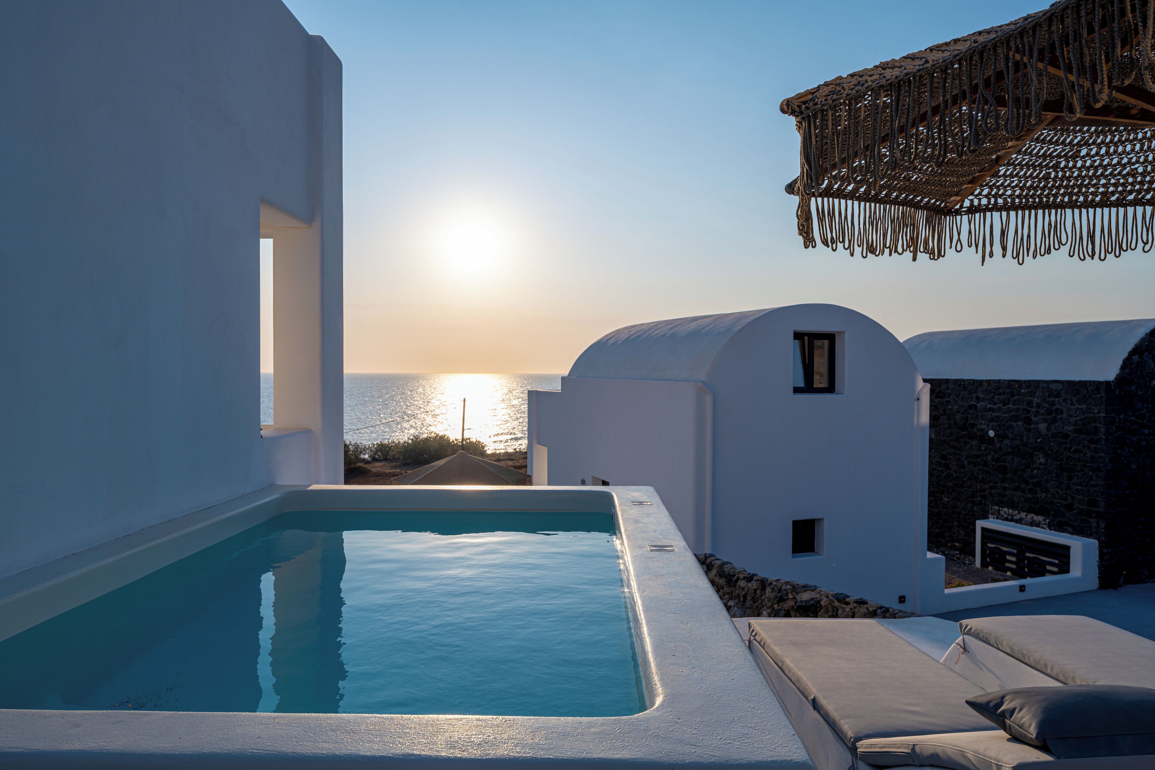 Zona de piscina con vistas al mar al atardecer, edificios blancos y un muro de piedra.