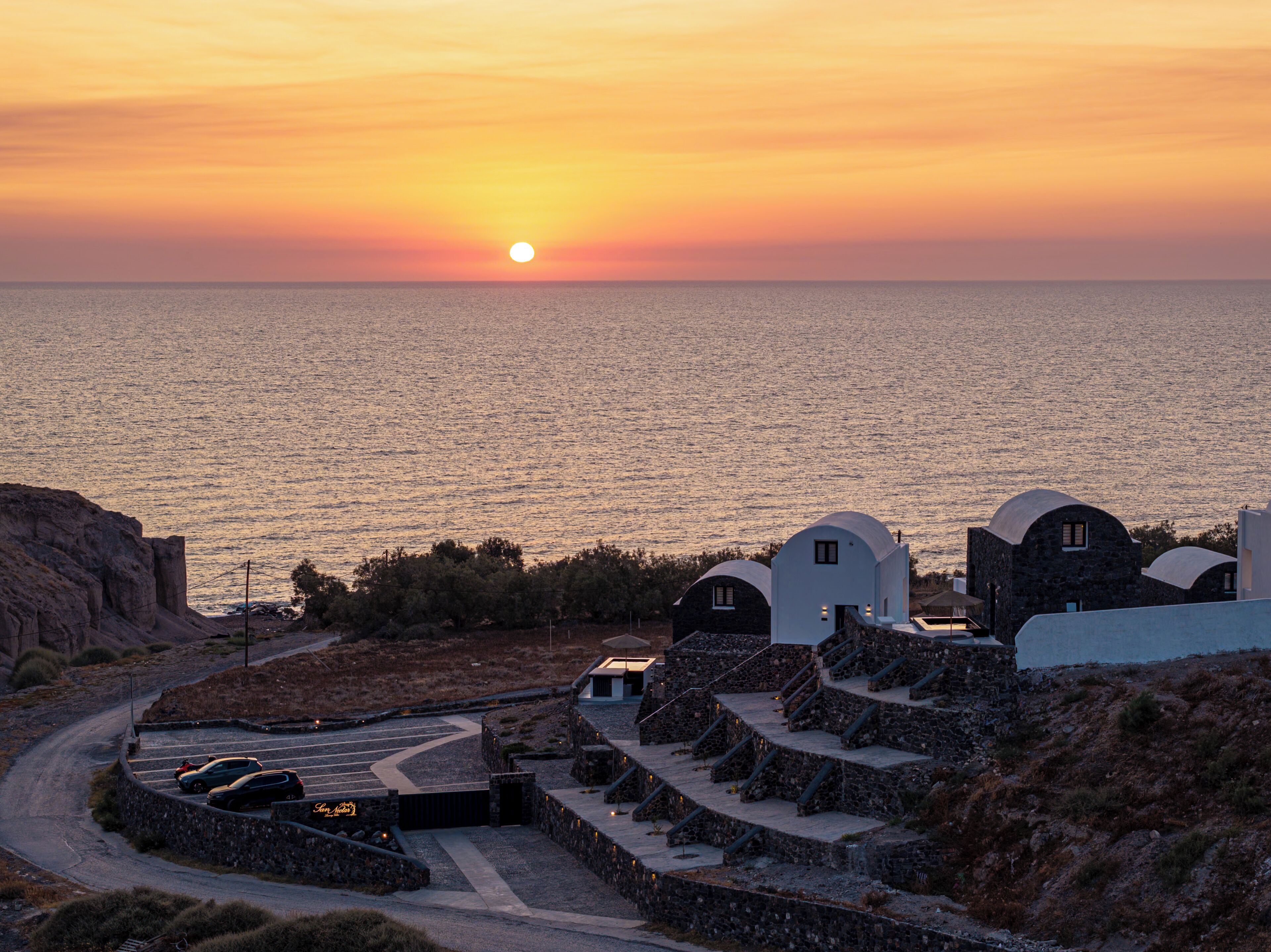 A coastal property with a curved driveway, stone steps leading to a white building with a dome, and a view of the sea at sunset.