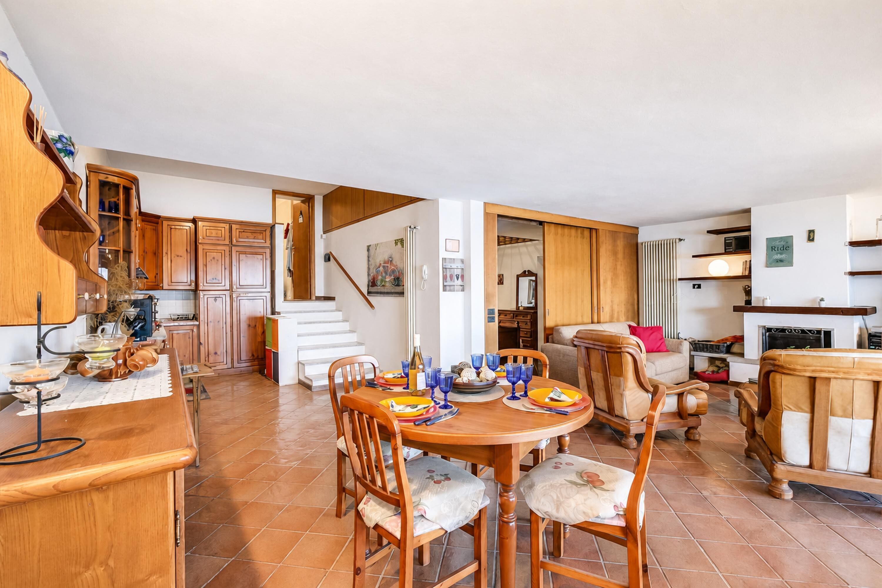 Un'ampia cucina con mobili in legno, una zona pranzo con un tavolo rotondo e sedie, e una scala che conduce a un piano superiore.