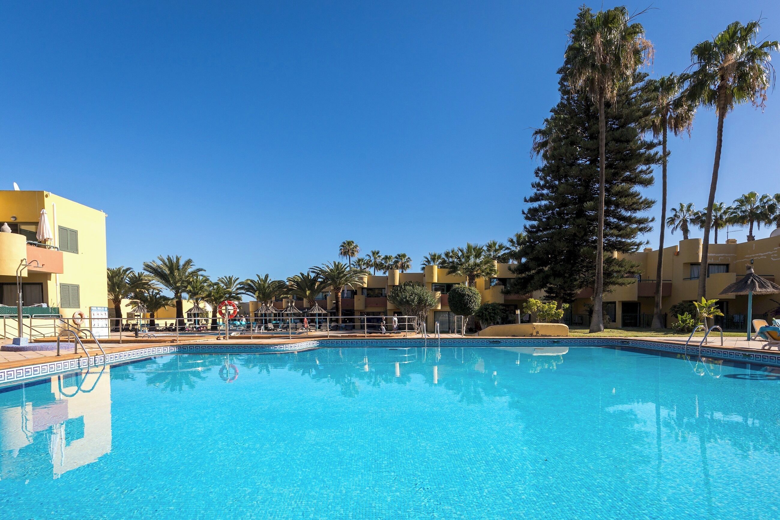 Piscina azul cristalina rodeada de palmeras y un edificio amarillo con una zona de estar cubierta.