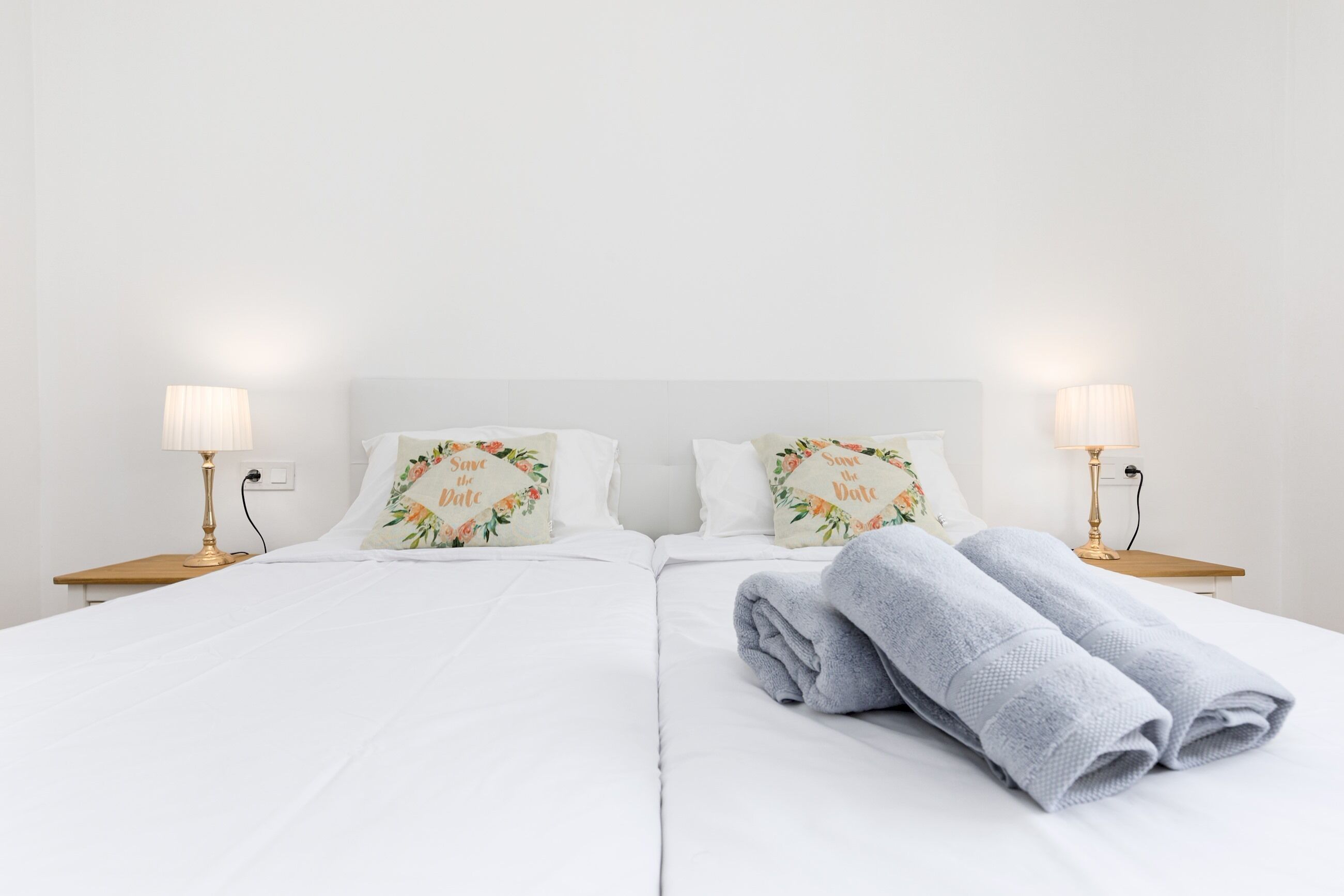 Un lit double avec une literie blanche, deux coussins décoratifs, deux lampes de chevet et une serviette pliée posée sur le lit.