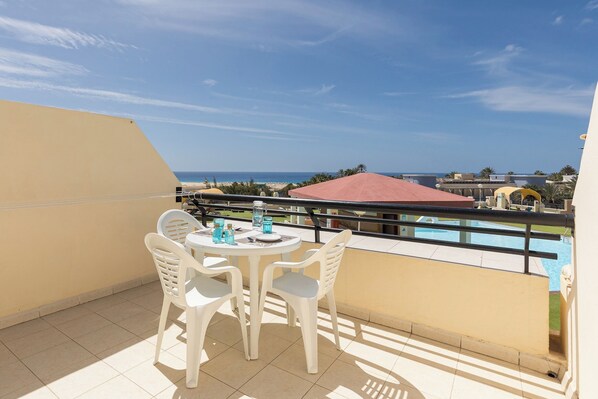 A balcony with a table and chairs, overlooking a pool and a beach.