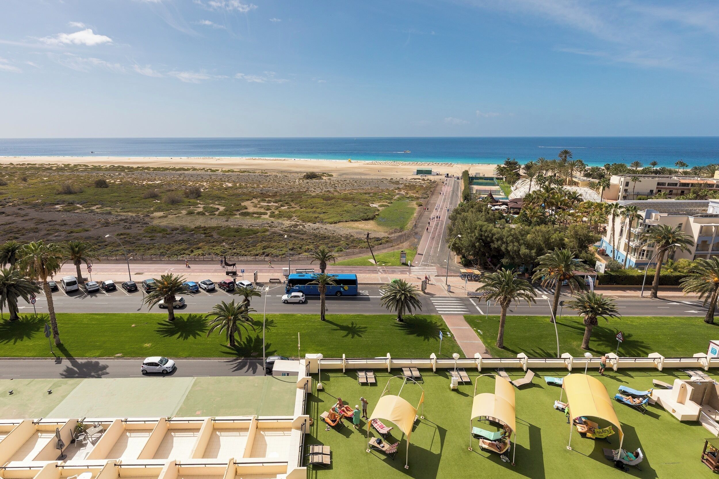 A coastal hotel with a view of the beach, a parking area, and outdoor seating with umbrellas.