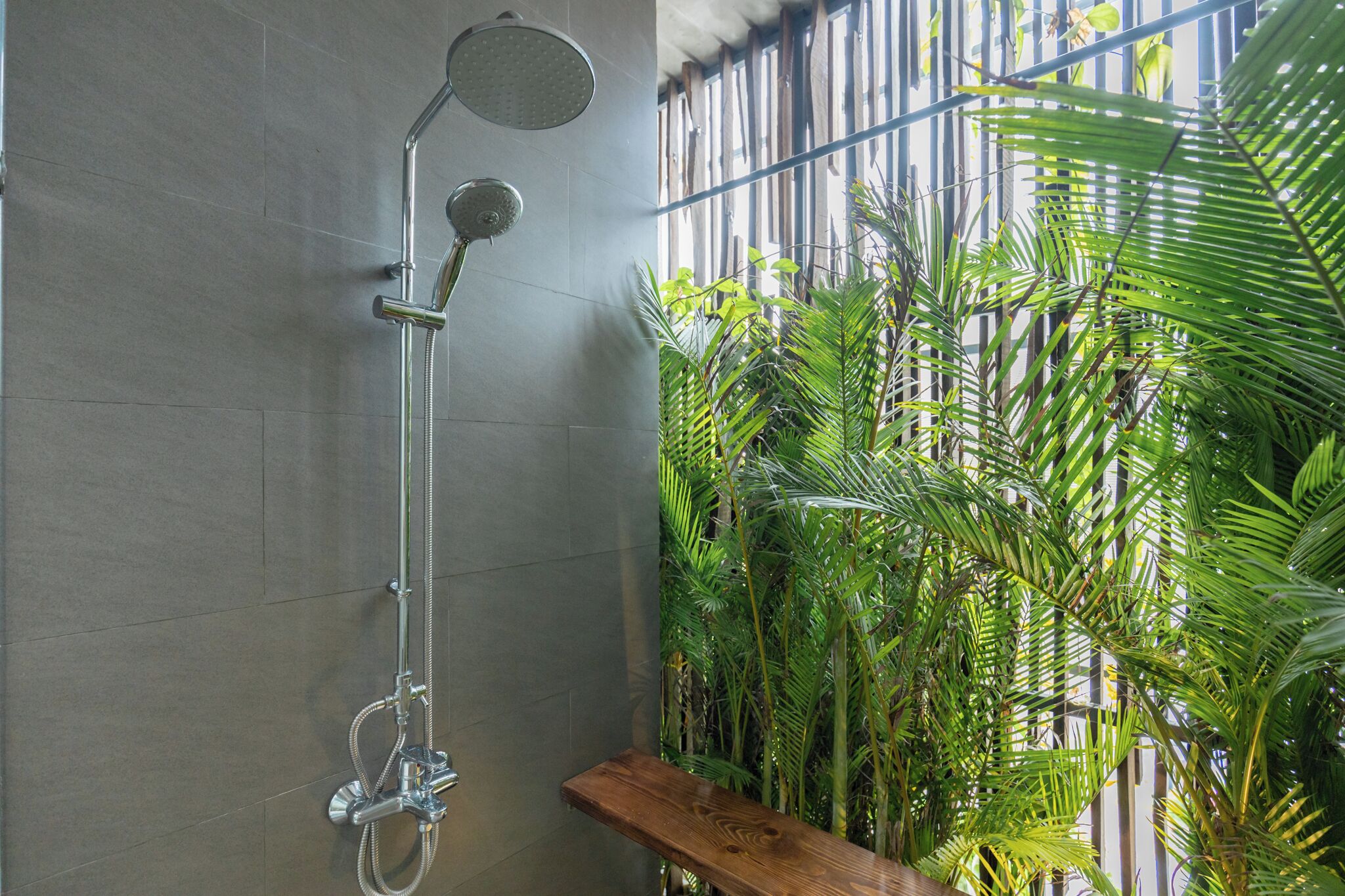 Une douche dotée d’un pommeau de douche à effet pluie et d’un pommeau de douche à main, d’un banc en bois et d’un mur agrémenté de stores verticaux et de plantes vertes luxuriantes.