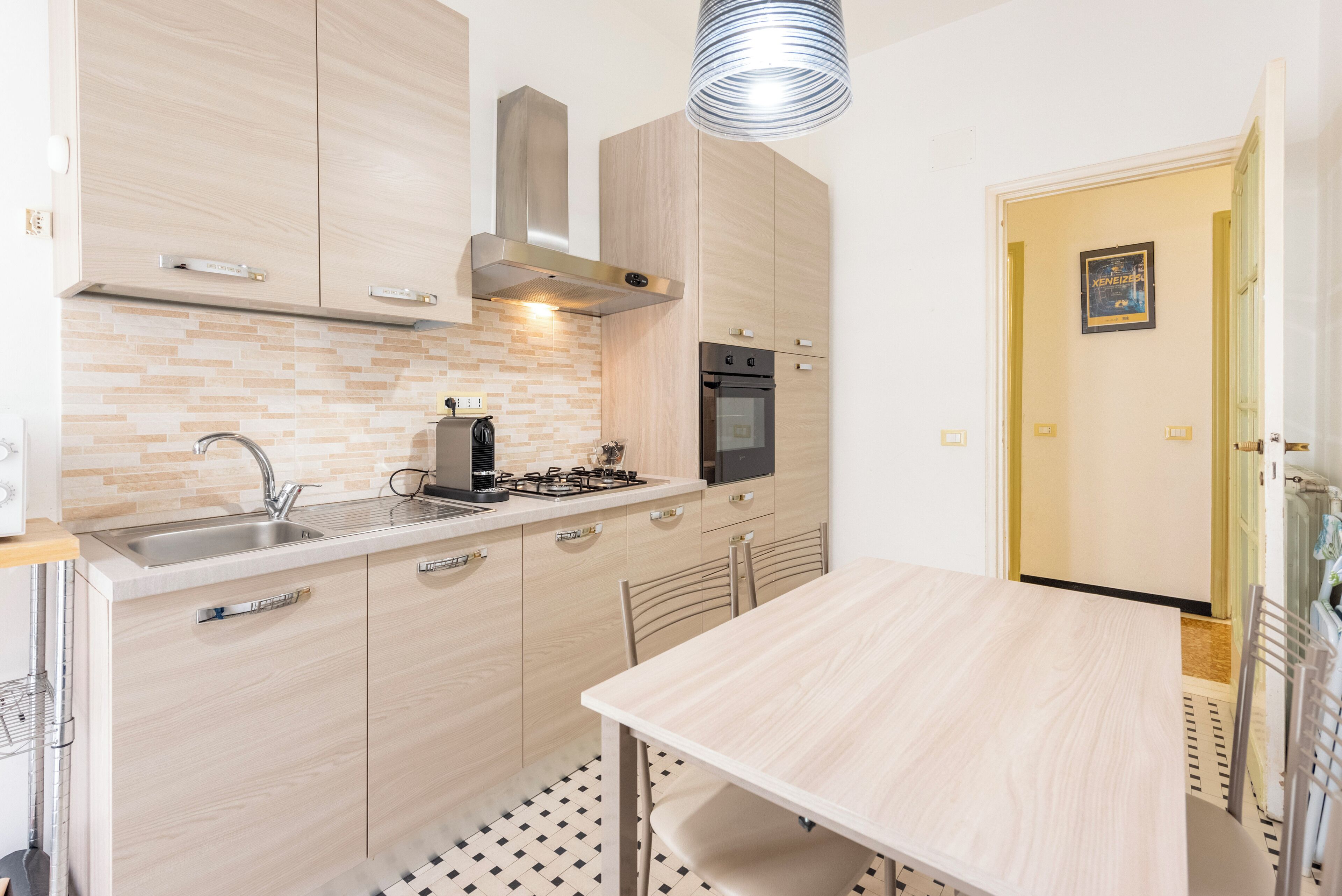 A modern kitchen with light wood cabinets, a stainless steel range hood, and a black oven. There's a dining table with chairs and a hanging light fixture.