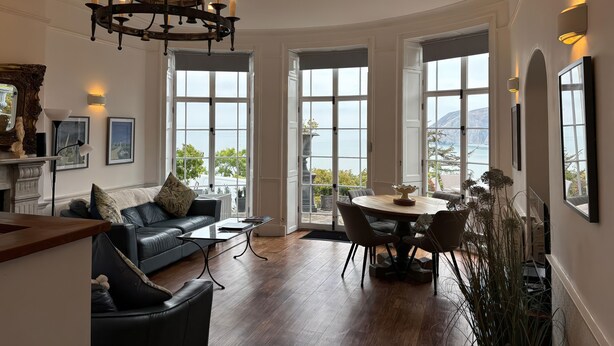 Een ruime woonkamer met een zwarte leren bank, een ronde eettafel met stoelen, een kroonluchter en grote ramen met uitzicht op zee.