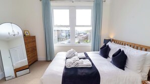 A neatly made bed with white and navy bedding, a wooden headboard, and a view of the outside through a window with light blue curtains. - Dinguardi (Seahouses)