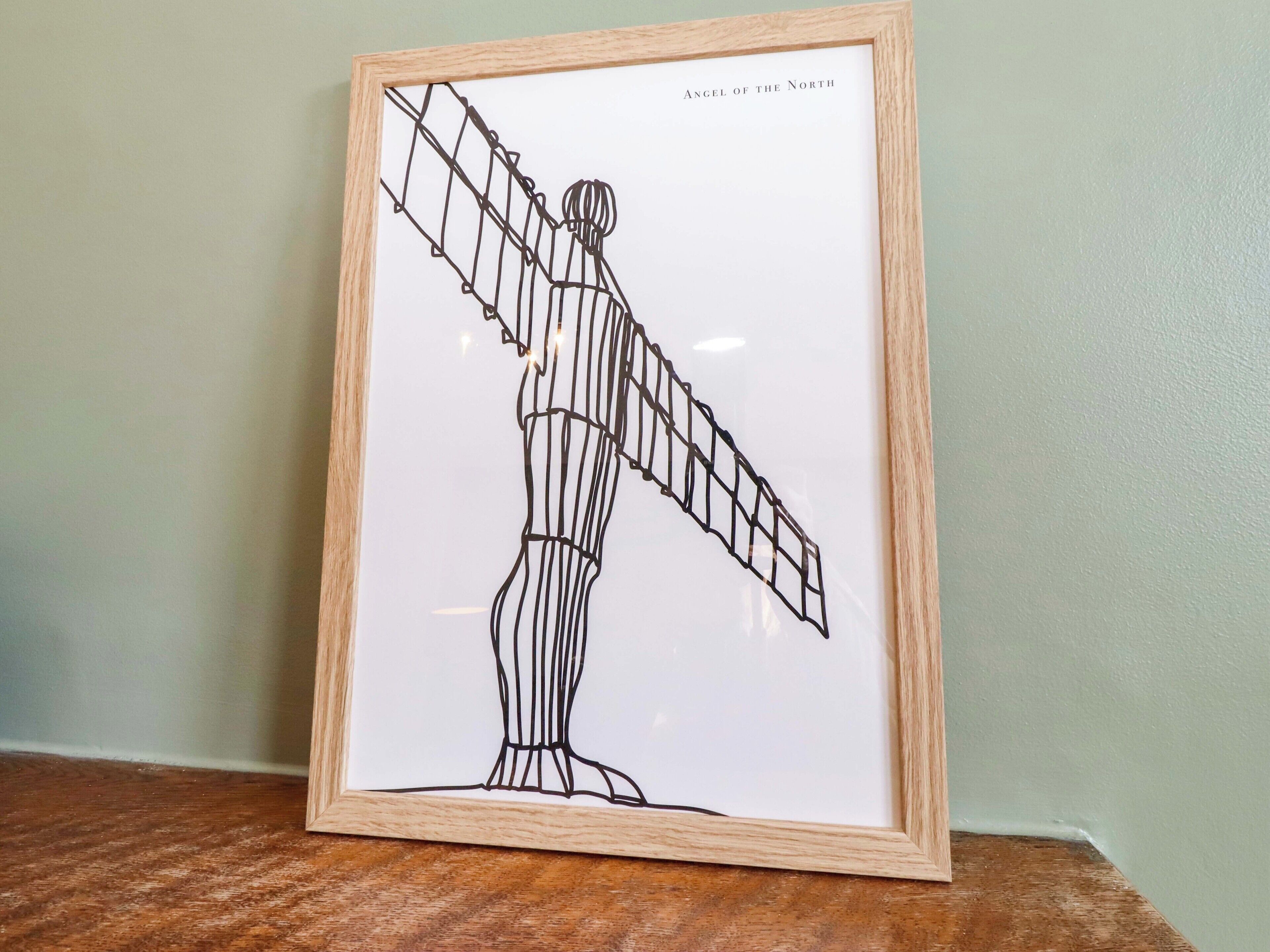 A framed artwork titled "Angel of the North" depicting a human figure with outstretched arms, standing on a base.