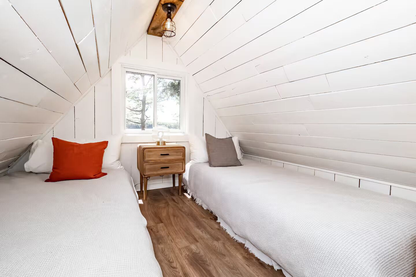 Ein kleines Schlafzimmer im Dachgeschoss mit weißen Paneelen, zwei Betten, einem Nachttisch und einem Fenster.