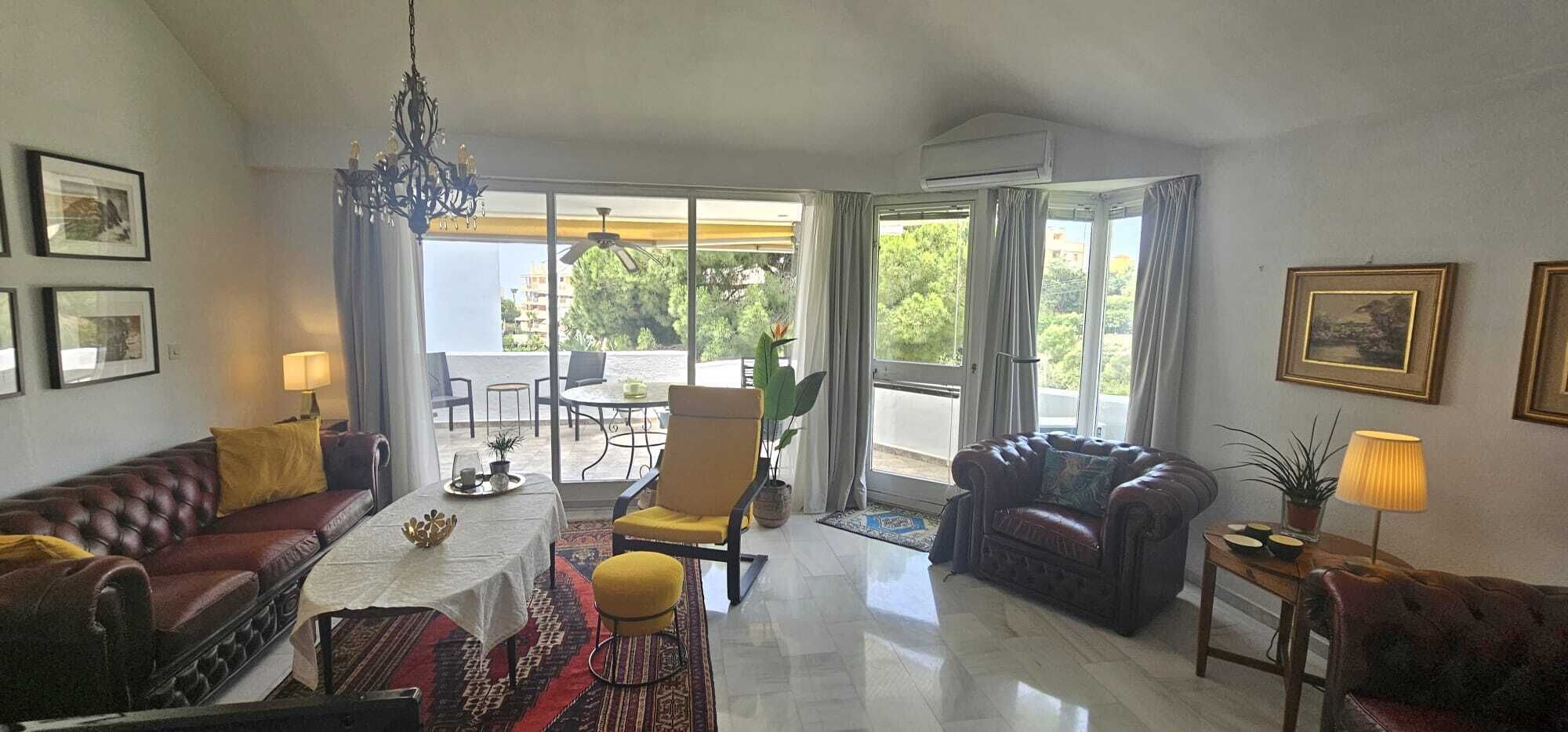 Ein großzügiger Wohnraum mit Sofa, Sessel und Esstisch. Ein Balkon bietet Blick auf Grünflächen und Gebäude.