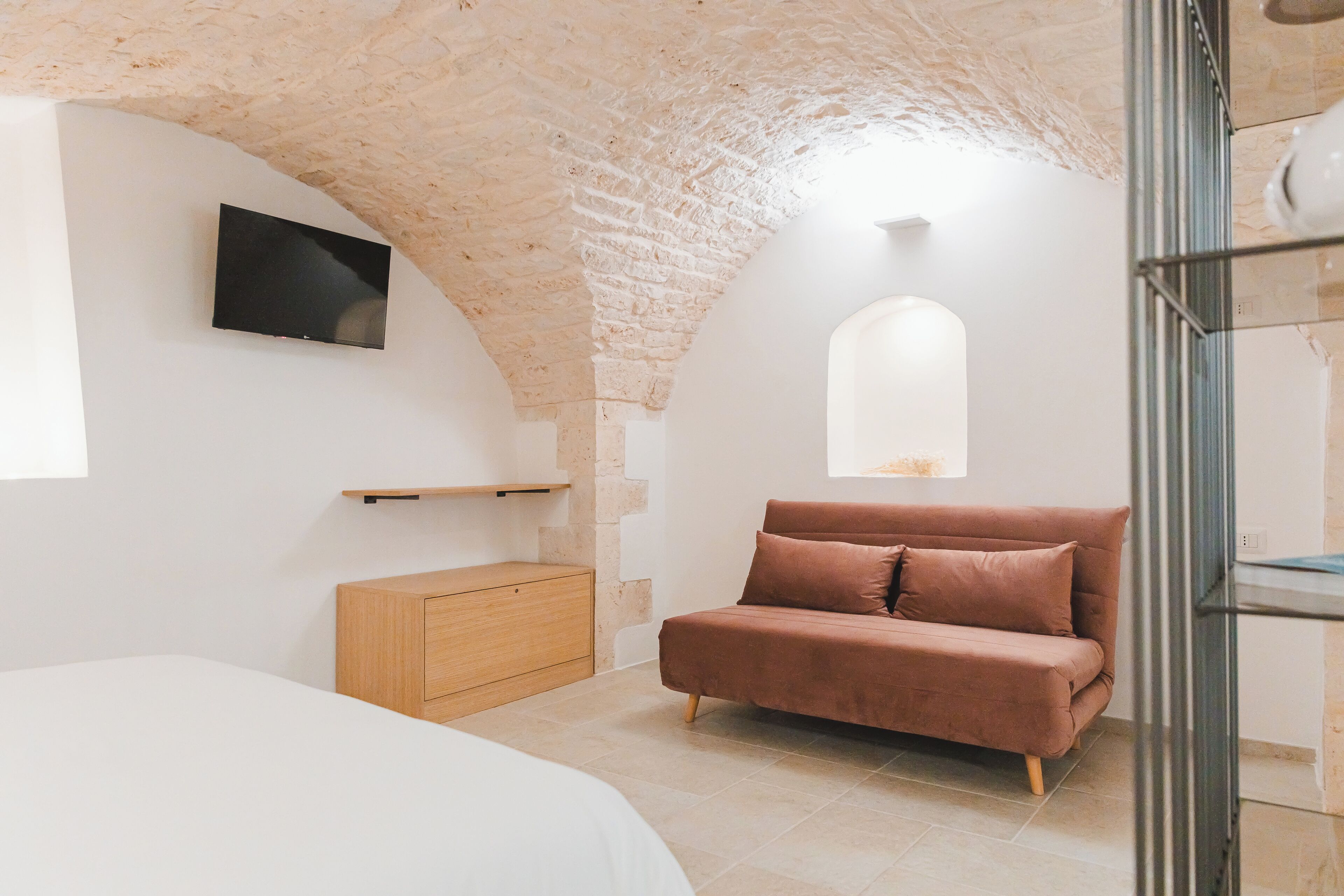 Een slaapkamer met een stenen plafond, een bed, een bruine bank, een houten dressoir en een televisie die aan de muur is bevestigd.
