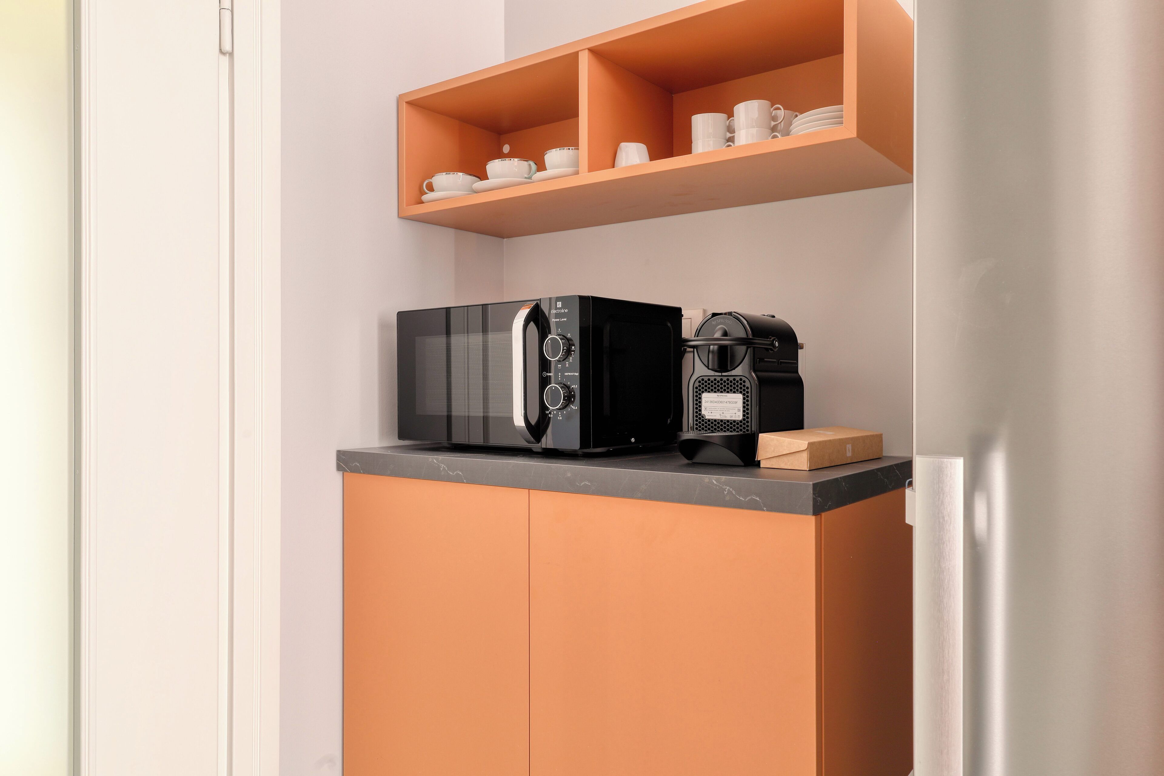A kitchen with a microwave, coffee maker, and a shelf with cups and plates.