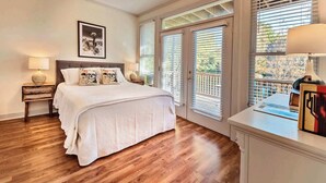 A bedroom with a large bed, wooden flooring, a ceiling fan, and a window with blinds.