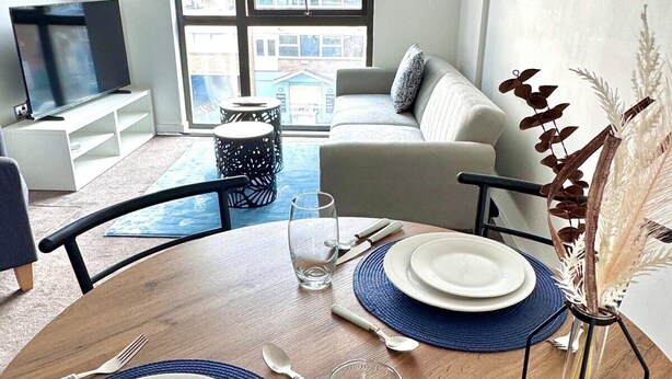 Un soggiorno moderno con zona pranzo, divano, televisore e vista sugli edifici attraverso ampie finestre.