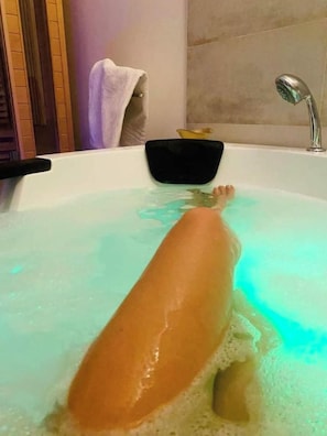 Une baignoire dans laquelle une personne est immergée, un objet noir posé sur le bord et une serviette accrochée au mur.