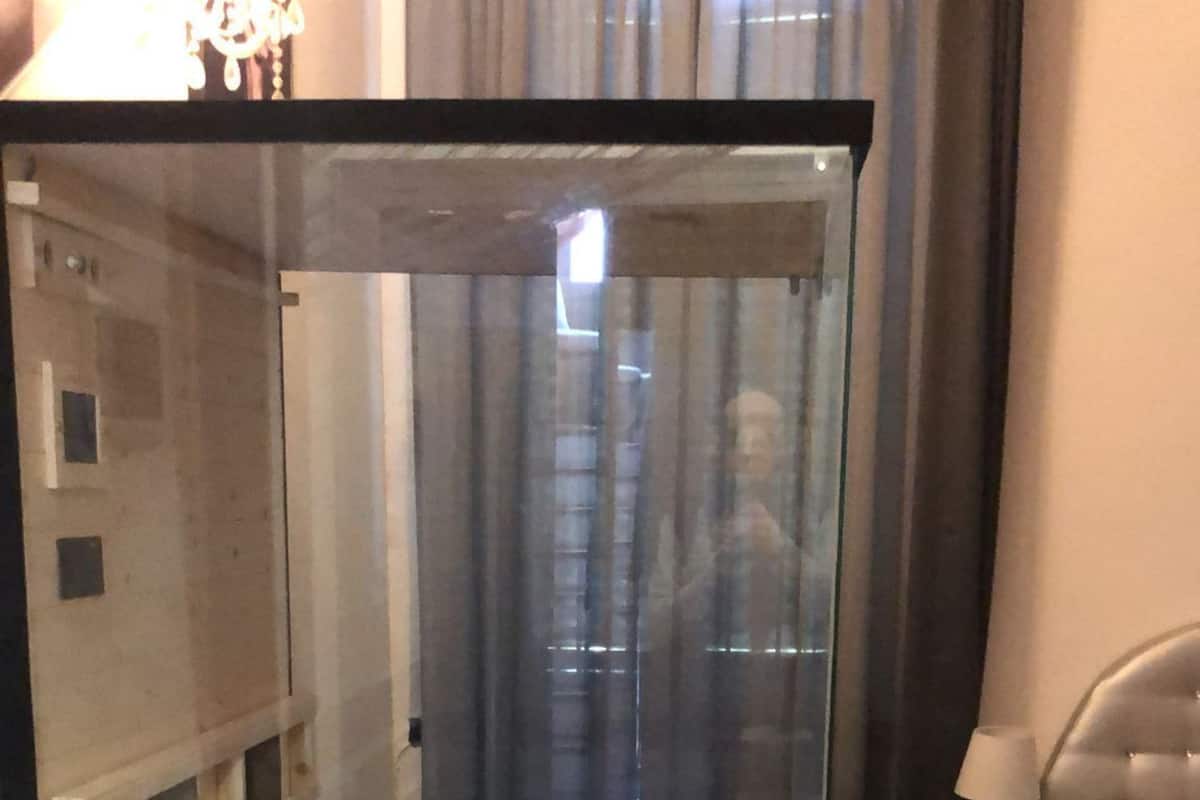 A room with a glass partition, a bed, a bedside table with a lamp, and a small wooden bench.