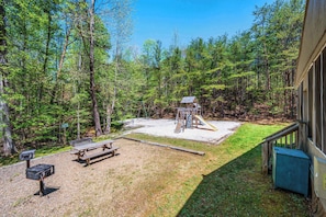 A backyard with a wooden deck, picnic table, grill, and playground equipment.