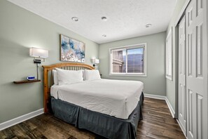 A bedroom with a bed, a nightstand, a wall-mounted lamp, a painting, and a window with blinds.