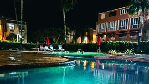 En oplyst swimmingpool om natten med liggestole og parasoller, omgivet af bygninger og grøn vegetation.