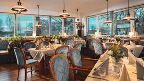 A restaurant with multiple tables and chairs, large windows, and hanging pendant lights. - Zur Rennbahn (Drage)