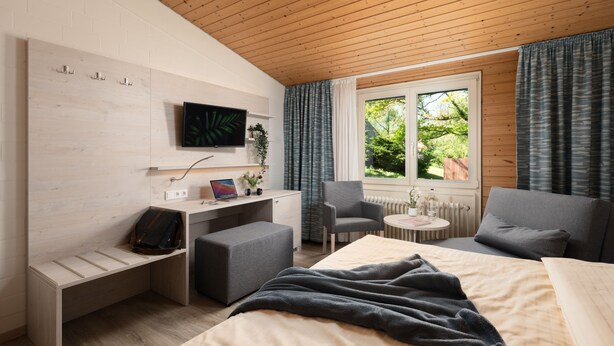 A modern hotel room with a wooden ceiling, a bed with a beige duvet, a grey sofa, a desk with a laptop, a chair, and a TV mounted on the wall. - Zur Rennbahn (Drage)