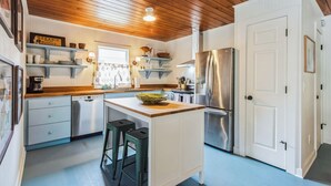 A modern kitchen with wooden ceiling, white cabinetry, stainless steel appliances, and a central island with stools. - Byrd Shack - Walk to Edisto Beach! (Edisto Island)