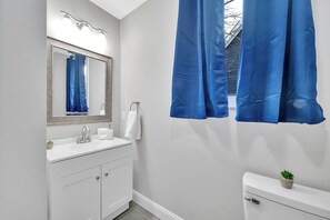 A bathroom with a white sink, a mirror with lights, a toilet, and blue curtains. - Charming 3BR Home, fenced yard, BBQ, mini golf, yard games--minutes to uptown (Charlotte)
