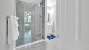 Casa de banho moderna com cabine de duche em vidro, azulejos brancos e tapete azul. É visível uma cama na divisão adjacente.