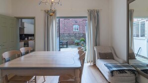 Ein Essbereich mit einem Holztisch und Stühlen, einer Couch mit kariertem Überwurf und Blick auf ein Backsteingebäude durch eine offene Tür.