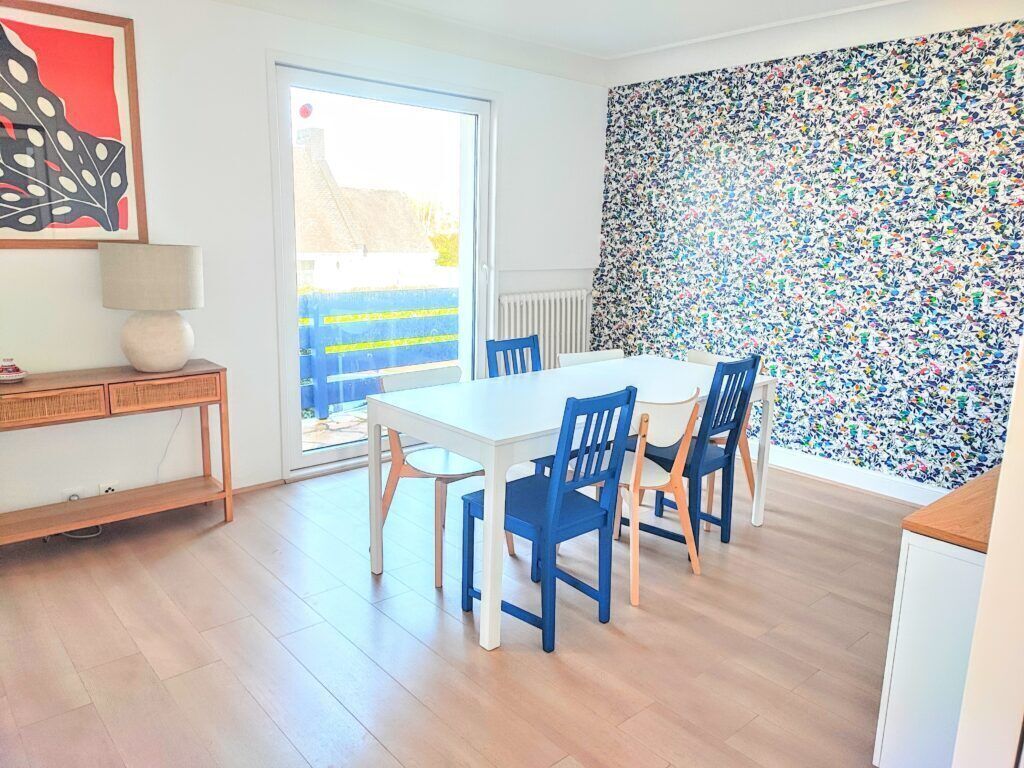 Un coin repas avec une table blanche et des chaises bleues, un plancher en bois, un radiateur et un mur décoré de motifs floraux colorés.