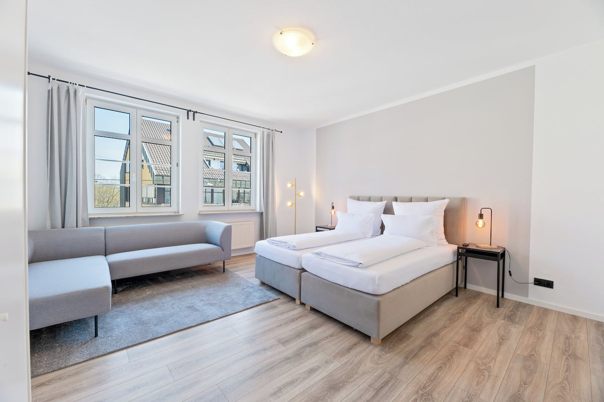 A modern hotel room with a large bed, a sofa, and a view of buildings outside the window.