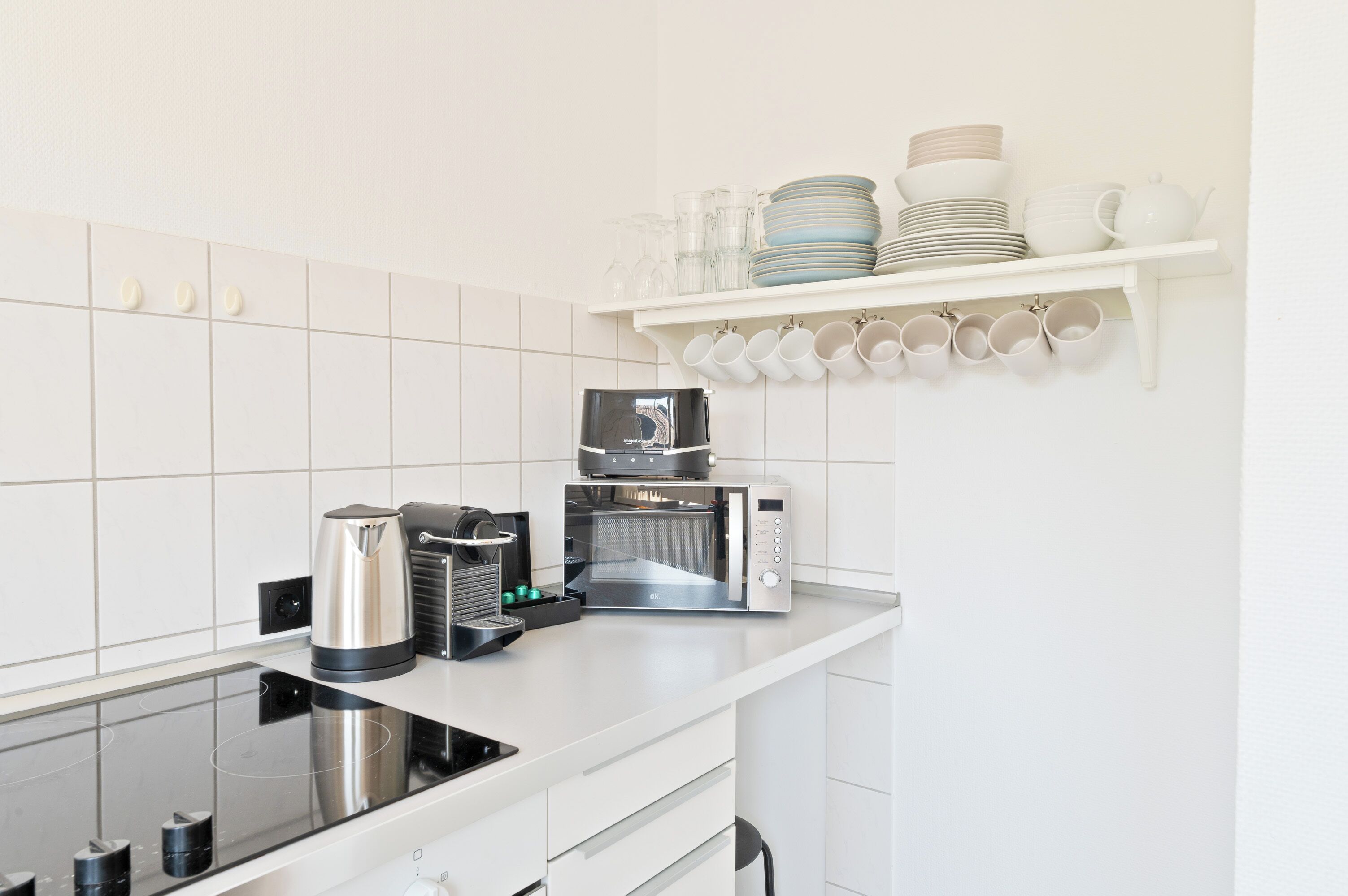 Une cuisine moderne équipée d’une cuisinière intégrée, d’une machine à café, d’un four à micro-ondes et d’une étagère où sont rangées assiettes et tasses.