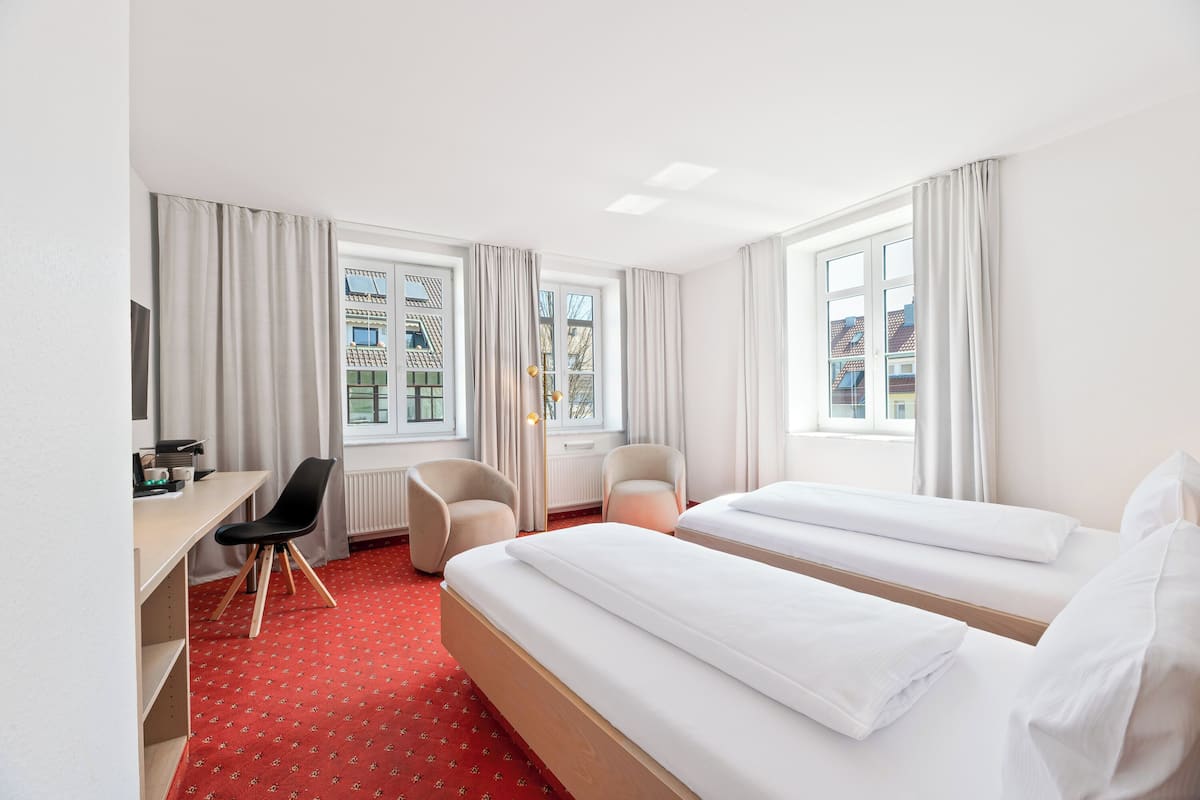 A hotel room with two beds, a desk, chairs, and large windows with curtains.
