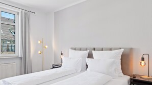 A double bed with white linens, a bedside table with a lamp, and a view of a building through a window with white curtains. - Steiners Hotel (Stuttgart)