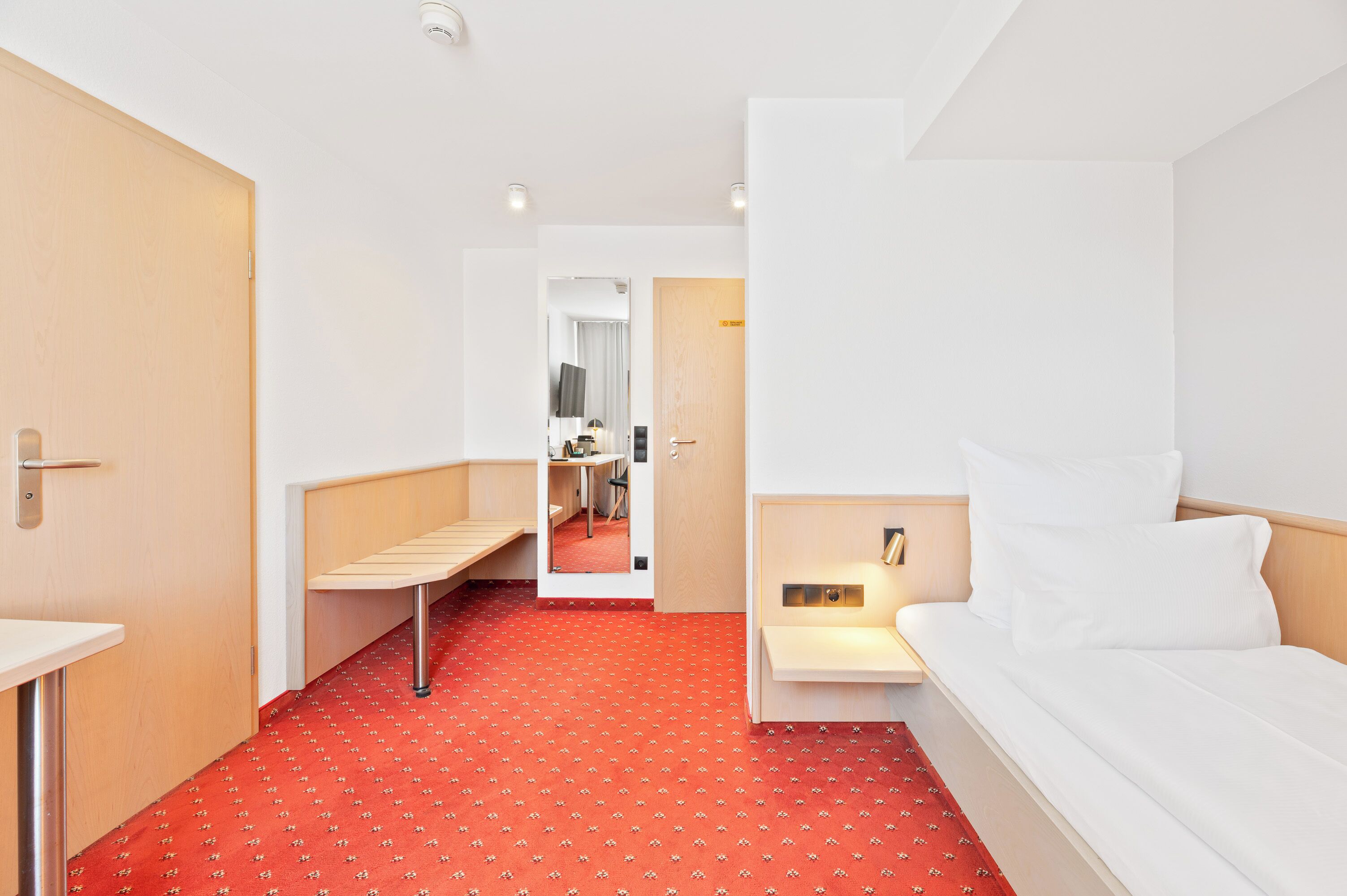 A hotel room with a red patterned carpet, a bed, a bedside table, and a small desk with a chair.
