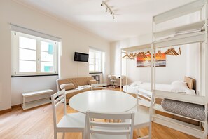 Et moderne, godt oplyst værelse med et rundt bord, hvide stole, en sofa, køjeseng og et tv på væggen.