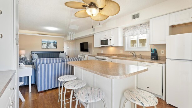 A compact kitchen with a microwave, sink, and white cabinets. There is a small island with stools and a ceiling fan. - Siesta Villa 104s (Ocean City)