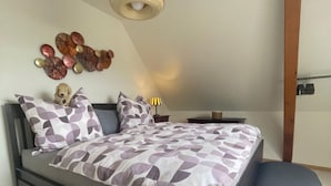 A modern bedroom with a bed, a bench, a wall-mounted lamp, and decorative wall art. - Le square Spacious apartment with terrace & garage, quiet location near Colmar  (Sundhoffen)