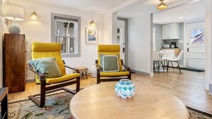 Un salón moderno con dos sillones amarillos, una mesa de centro redonda de madera y un jarrón. Se ve una cocina al fondo.