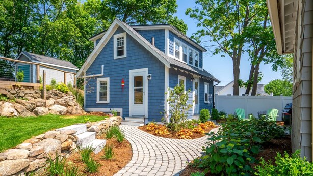 Una casa de dos pisos con fachada azul, un muro de piedra de contención y un camino pavimentado que conduce a la puerta principal.