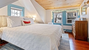 Un dormitorio con techo inclinado, una cama con ropa de cama a rayas, una mesita de noche, un aparador y una ventana con vistas al exterior.