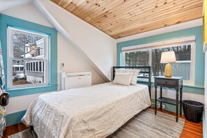 A bedroom with a bed, a nightstand, a lamp, and a window with a view of a house.