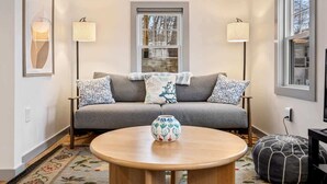Una sala de estar acogedora con un sofá gris, cojines estampados, una mesa de centro redonda de madera y dos lámparas de pie.