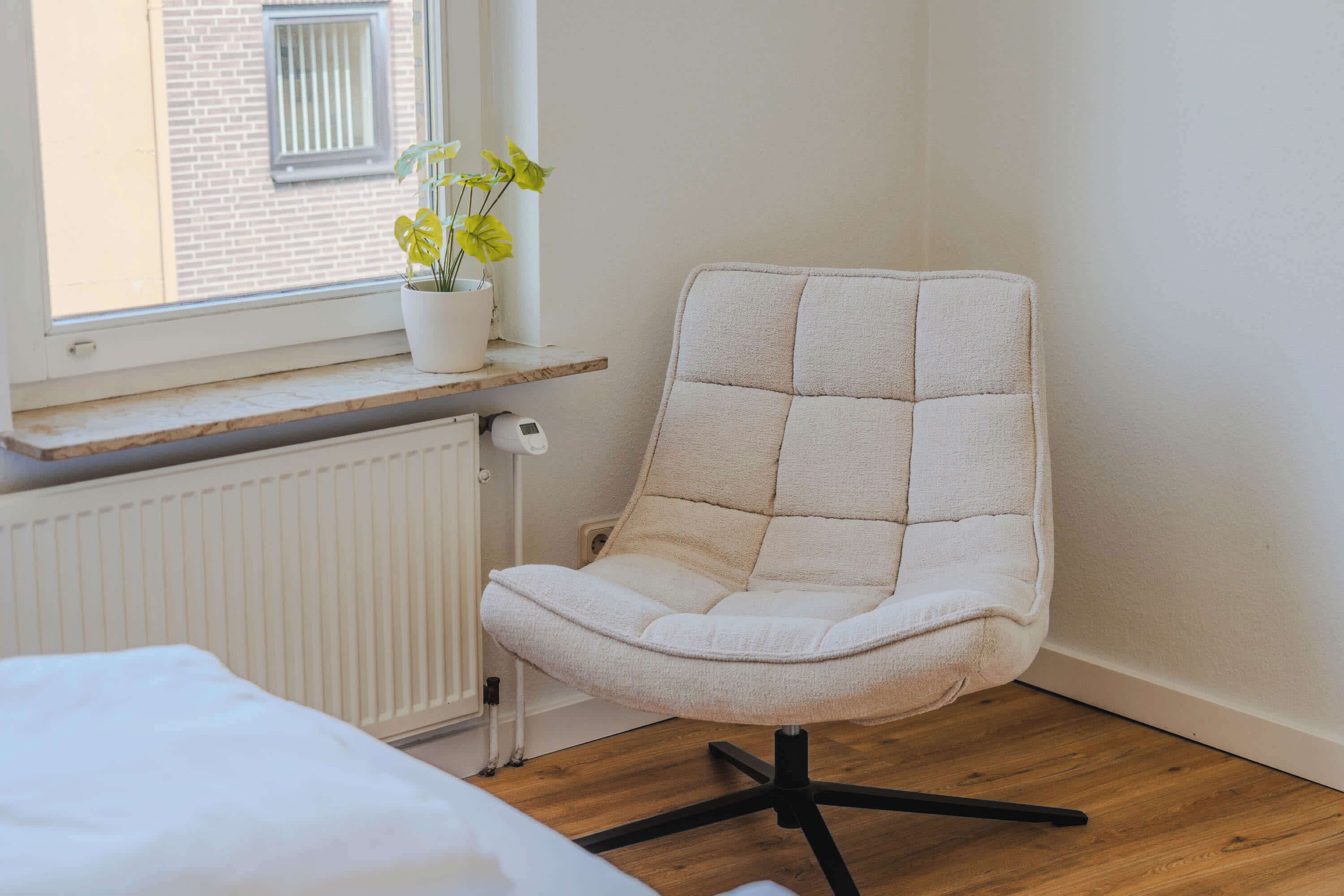 Une pièce avec une chaise blanche, un plancher en bois, un radiateur et une fenêtre donnant sur un immeuble en briques.