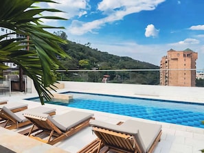 A swimming pool with lounge chairs, a clear sky, and a hill with buildings in the background. - H105 Design & Comfort | Heart of Granada | Pool (Cali)