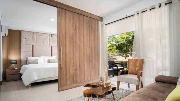A modern bedroom with a large bed, wooden panel wall, and a balcony with outdoor furniture. - H108 | 2-Bed, 2-Bath  | Pool | Gym | 24/7 Security (Cali)