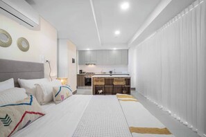 Quarto de hotel moderno com uma cama, mesa de cabeceira e uma cozinha compacta ao fundo.