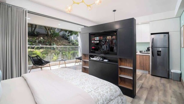 A modern bedroom with a large bed, a TV mounted on a wall, a balcony with a chair, and a kitchen area with a refrigerator. - H401 | top-Rated Cali Retreat 2BR w/ Balcony & Pool 24/7 Security + Free Parking (Cali)