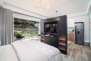 Un dormitorio moderno con una cama grande, un televisor montado en la pared, un balcón con una silla y una zona de cocina con un refrigerador.